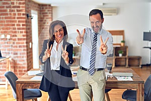 Two middle age business workers standing working together in a meeting at the office smiling looking to the camera showing fingers