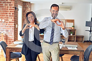 Two middle age business workers standing working together in a meeting at the office smiling in love doing heart symbol shape with