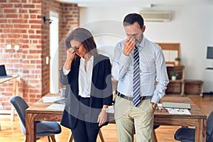 Two middle age business workers standing working together in a meeting at the office tired rubbing nose and eyes feeling fatigue