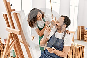 Two middle age students smiling happy playing with paintbrush painting to each other at art school