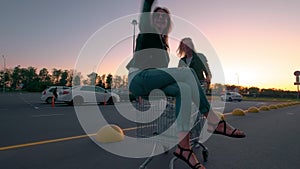 Two millennials girlfriends in street clothes have fun in a supermarket parking lot at sunset. Riding a shopping cart