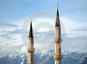 Two minarets rise against the backdrop of impregnable snow-capped mountains covered with trees under serene blue sky