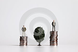 Two miniature men standing on a pile of coins on either side of a tree.