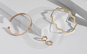 Two modern geometric shape golden bracelets and earrings pair on white podium with copy space