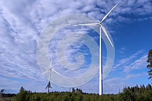 Two modern white windmills stand high above green forest, sunny summer day. Sweden, green energy