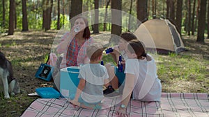 Two moms and two sons drinking cold beverages from fridge on picnic blanket during summer family camping vacation with