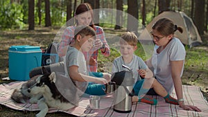 Two moms and two sons drinking tea on picnic blanket during summer family camping vacation with tent in forest. Boy