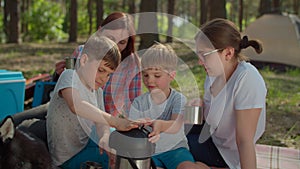 Two moms and two sons drinking tea on picnic blanket during summer family camping vacation with tent in forest. Boy