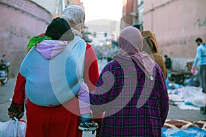 Two moroccan women with child in sling seen from the back dressed in typical blue and brown djellaba walk down the