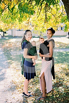 Two mothers with babies in slings stand under a tree in the park