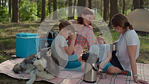 Two mothers and two sons drinking tea on picnic blanket during summer family camping vacation with tent in forest. Happy