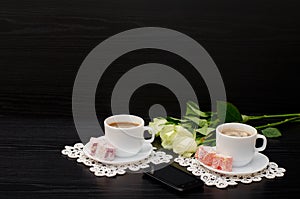 Two mugs of coffee with milk, smartphone, Turkish delight on a saucer, white roses on a black background.