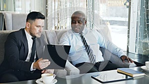 Two multi-ethnic businessmen looking at clipboard and discussing startup projects in glassy cafe during lunch time