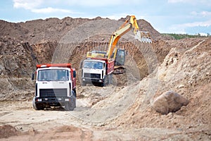 Two multi-ton mining trucks in process of loading ore by orange excavator for exporting minerals from the open-cast mine quarry