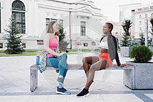 Two multiethnic curvy girls relaxing after exercise session, outdoor communicating.
