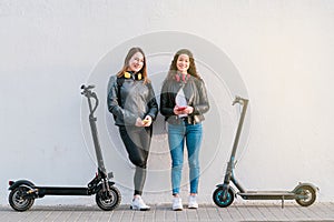 Two multiethnic female friends on electric scooters using smartphone outdoors.