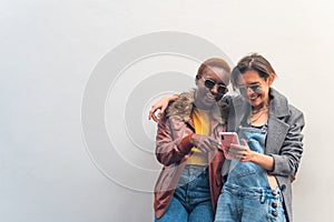 two multiethnic friends looking a message on a smart phone in the street and smiling with a white wall in the background