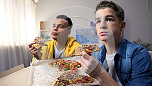 Two multiethnic male teenagers eating pizza and watching TV show, leisure time