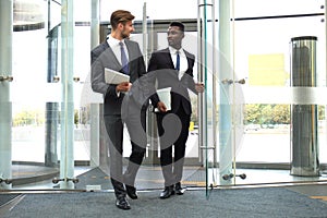 Two multinational young businessmen entering in office building with glass doors.