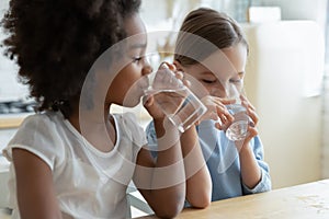 Two multiracial girls sit in kitchen feels thirsty drink water