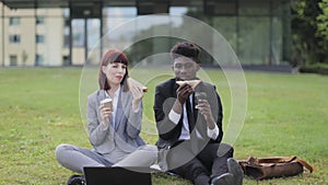 Two multiracial successful partners sitting on green grass outside modern office, enjoying their lunch