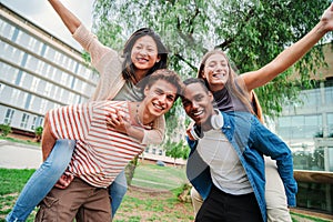 Two multiracial teenage student best friends giving a piggyback ride to their girlfriends. Young men smiling and having