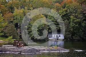 Two Muskoka chairs sitting on a rocky shore facing a calm lake with trees and a white cottage in the background