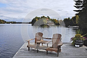 Two Muskoka chairs sitting on a wood dock facing a calm lake. Across the water is a cottage nestled among green trees