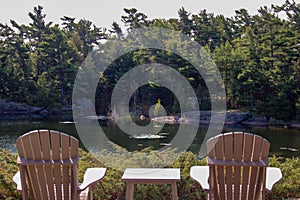 Two Muskoka chairs sitting on a wood dock facing a calm lake. Across the water is a white cottage nestled among green trees