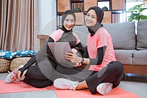 Two Muslim women wearing hijab sportswear smile while relaxing on the floor while using a digital tablet