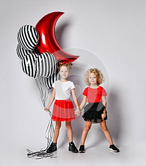 Two nice kids girls in skirts and t-shirts are standing holding hands, ready for a party with stylish air balloons