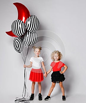 Two nice smiling kids girls in skirts and t-shirts are standing holding hands and with air balloons