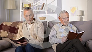 Two offended male friends reading books on sofa at home, misunderstanding