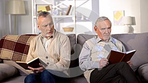 Two offended male friends reading books on sofa home, misunderstanding conflict