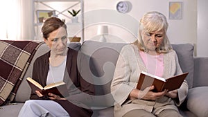 Two offended old women reading books sitting nursing home sofa, misunderstanding