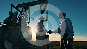 Two oil workers talking and shaking hands with pumpjack working in background