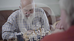 Two old men playing chess sitting at home on the leather sofa. Bearded man thinking which chess piece to make a move