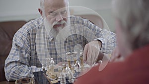 Two old people playing a game of chess sitting at home on the leather sofa. Bearded man making a move. Caucasian old men