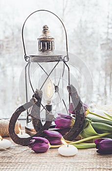 Two old rusty horseshoes, brown vintage lantern with modern dimmable decorative light bulb switched on and white spa candles lit a