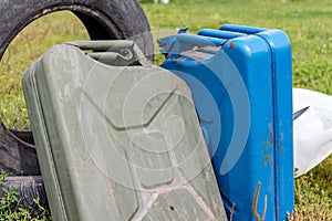 Two old and rusty metal military gasoline canisters with petrol on the grass and automobile tire behind
