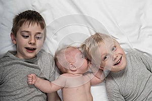 Two older brothers play and laugh with the youngest baby in a white bed. Happy childhood, big family