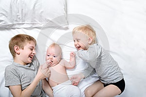 Two older brothers play and laugh with the youngest baby in a white bed. Happy childhood, big family