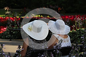 Two older women wearing wide white hats sitting together in a spa park full of colorful blooming flowers