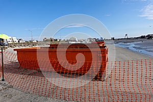 Two orange dumpsters at a construction site on a wet sandy beach near an orange mesh fence