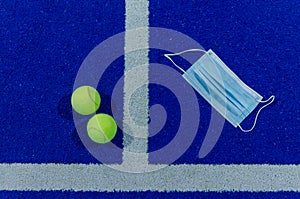 Two paddle tennis balls and a surgical mask next to the service lines of a blue paddle tennis court