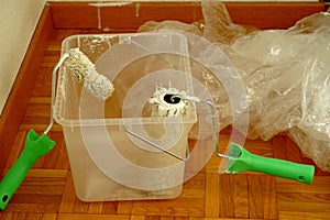 Two paint rollers in a container on the floor. The paint is dripping off the rollers. In the background there is a plastic foil to