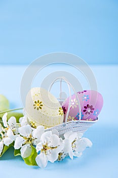 Two painted yellow and purple Easter eggs in basket with white spring cherry flowers on light blue background