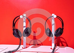 Two pairs of black headphones on stand. Red background, white table and black noise reduction device create creative atmosphere.