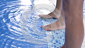 Two pairs of female legs stand on the side of the pool with blue clear water