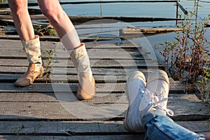 Two pairs of female legs on the wooden pier by the river. One girl in rolled-up blue jeans and white sneakers, another one in cowb